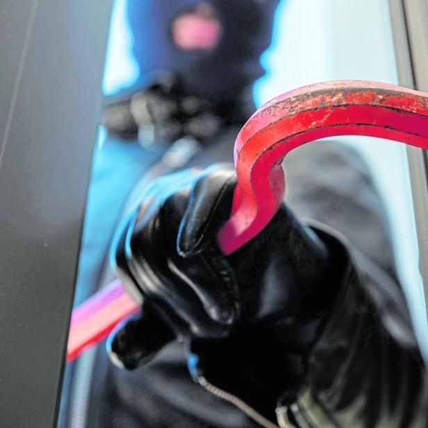 Eine vermummte Person mit Handschuhen hebelt mit einem roten Brecheisen ein Fenster auf.