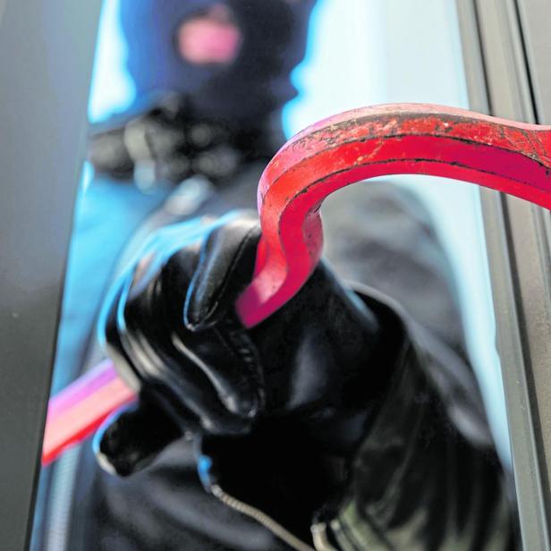 Eine vermummte Person mit Handschuhen hebelt mit einem roten Brecheisen ein Fenster auf.