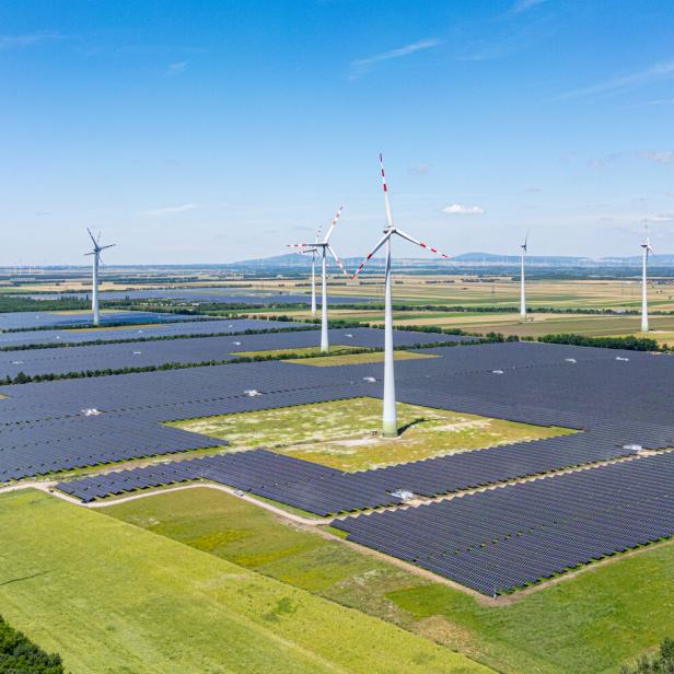 Mehrere Windräder und große Solarfelder stehen auf einer weitläufigen grünen Landschaft unter blauem Himmel.