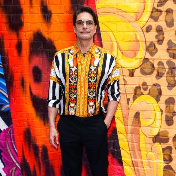 Ein Mann mit gemustertem Hemd und schwarzer Hose steht vor einer bunt bemalten Wand.