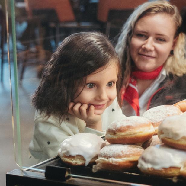 Eine erwachsene Frau kniet neben einem kleinen Mädchen, das in eine Vitrine mit Krapfen hineinschaut.