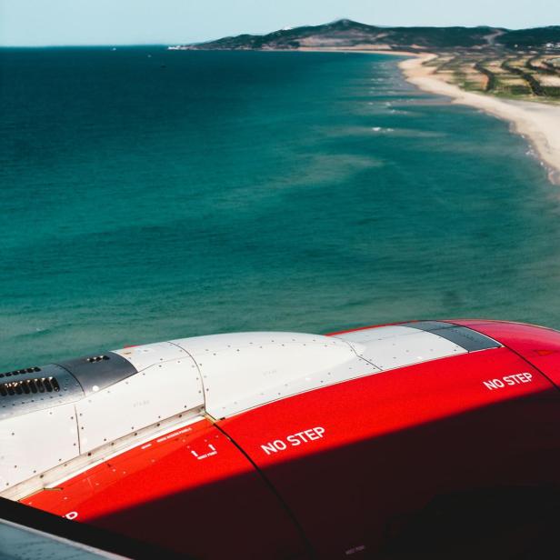 Blick aus einem Flugzeugfenster auf die Tragfläche mit der Aufschrift 'NO STEP' und eine Küstenlinie mit Strand und Meer im Hintergrund.