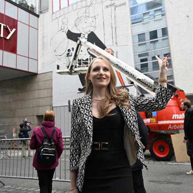 Eine Frau steht vor einer bemalten Hauswand mit der Aufschrift „Lugner City“ und zeigt auf das Wandbild.