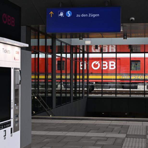 Ein roter ÖBB-Zug steht hinter Glaswänden, daneben ein Fahrkartenautomat und ein Wegweiser zu den Zügen.