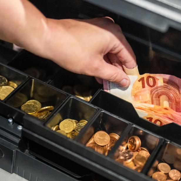 Eine Hand legt einen Zehn-Euro-Schein in eine geöffnete Ladenkasse mit Münzfächern.