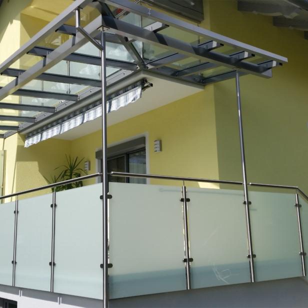 Balkon mit Edelstahlgeländer und Glasüberdachung an einem gelb gestrichenen Wohnhaus.