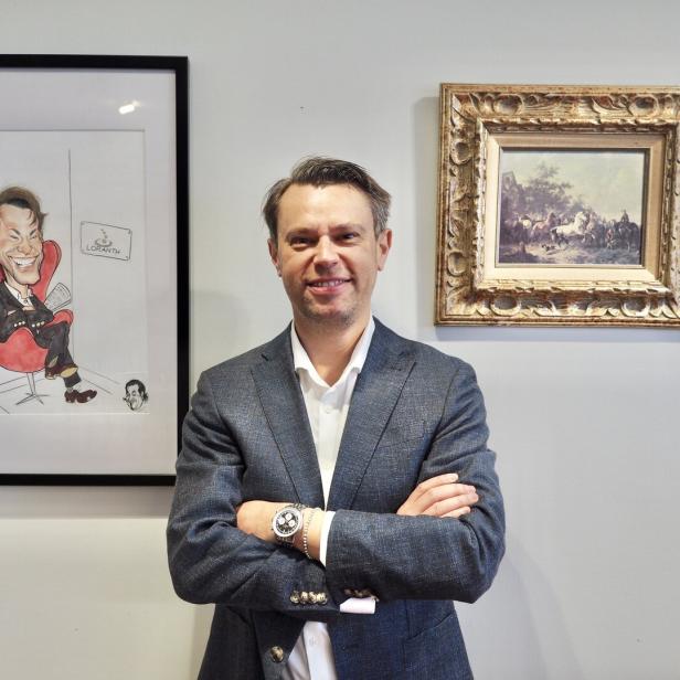 Ein Mann im Anzug steht lächelnd mit verschränkten Armen vor zwei gerahmten Bildern an einer Wand.