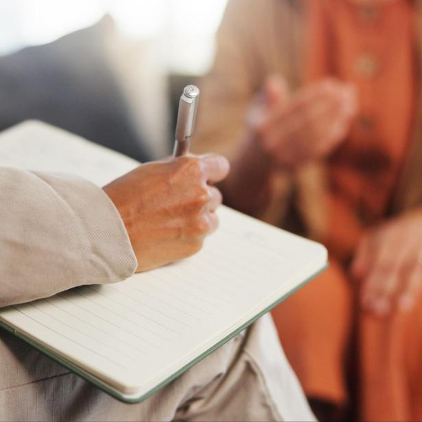 Ein Therapeut schreibt mit einem Stift in ein Notizbuch, die andere Person gestikuliert mit den Händen.