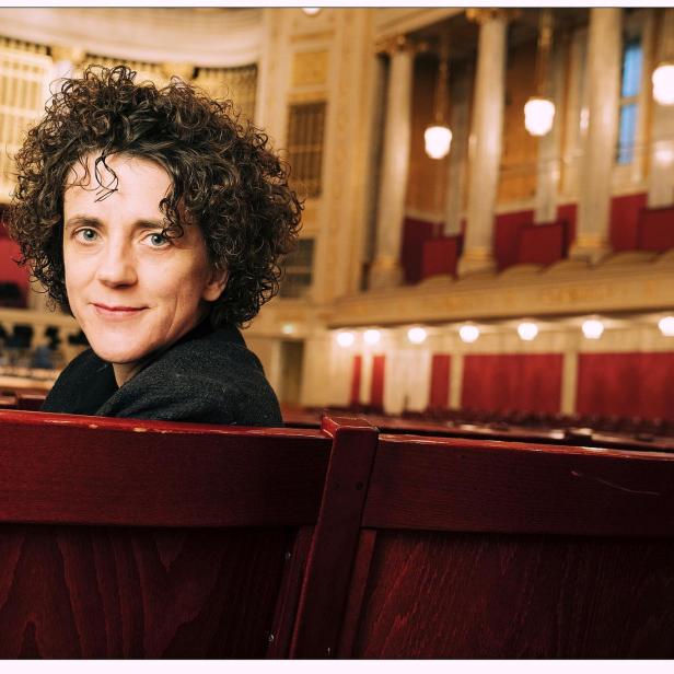 Eine Person mit lockigen Haaren - Olga Neuwirth - sitzt in einem eleganten Konzertsaal auf einer roten Holzbank und blickt lächelnd zurück.