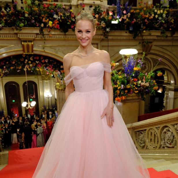 Eine Frau in einem eleganten, rosafarbenen Ballkleid steht auf einem roten Teppich vor festlich geschmückter Kulisse.