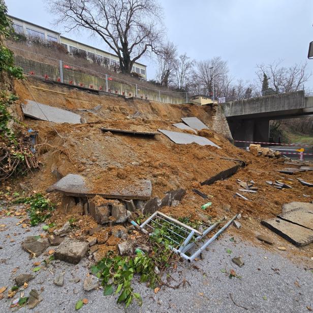 Eingestürzte Stützmauer mit Erdrutsch und Absperrungen im Bereich eines Wohngebäudes.