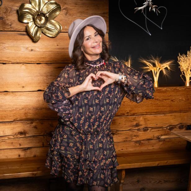 Frau mit Hut formt ein Herz mit den Händen vor einer Holzwand mit Dekoration.