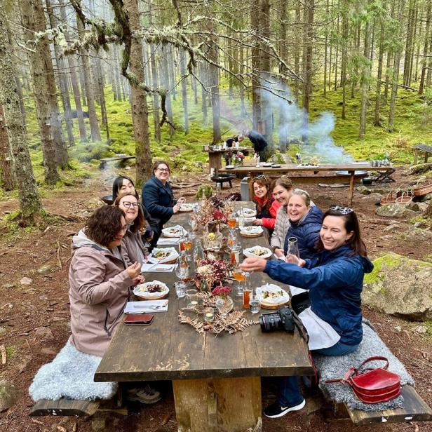 Eine Gruppe von Menschen sitzt an einem rustikalen Holztisch im schwedischen Wald und genießt gemeinsam ein Essen.