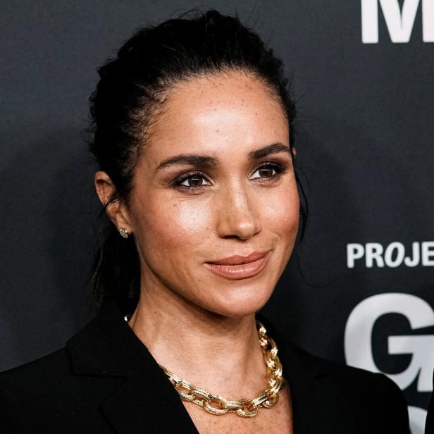 Herzogin Meghan mit hochgesteckten Haaren und goldener Kette vor dunklem Hintergrund.