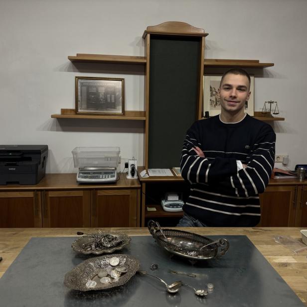 Ein junger Mann steht mit verschränkten Armen hinter einem Schreibtisch mit Silbermünzen und Schalen in einem Büro.