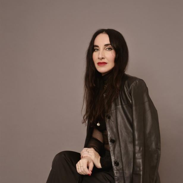 Frau -Mirna Funk -  mit langen dunklen Haaren trägt schwarze Lederjacke und schwarze Hose mit weißem Streifen, sitzt vor grauem Hintergrund.