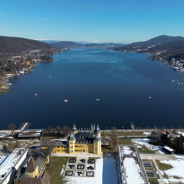 Ein gelbes Schloss liegt an einem See, umgeben von Bergen und Gebäuden.