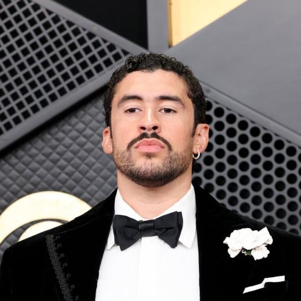 Bad Bunny mit Ohrring, Bart und Fliege trägt einen schwarzen Anzug mit weißem Hemd und Blume.