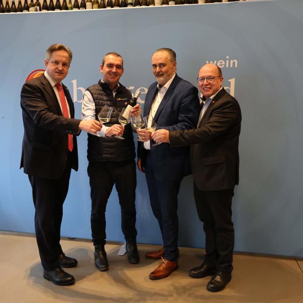 Vier Männer stoßen mit Weingläsern an und stehen vor einer blauen Wand mit Flaschen auf einem Regal darüber.