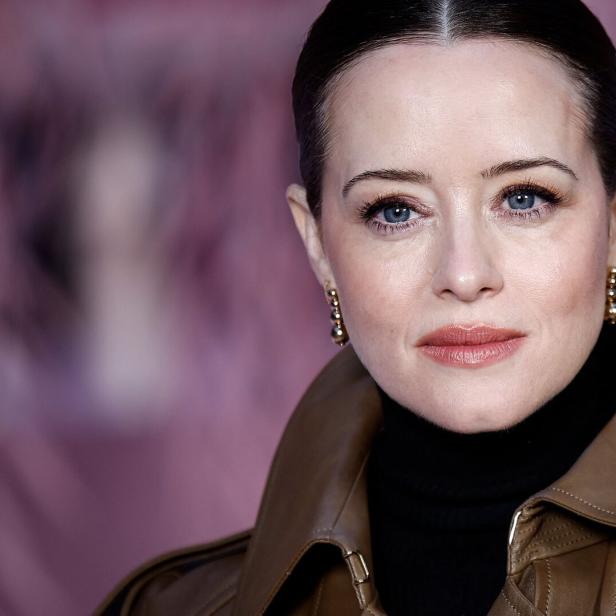 Claire Foy trägt einen braunen Ledermantel und Ohrringe vor einem verschwommenen Hintergrund.