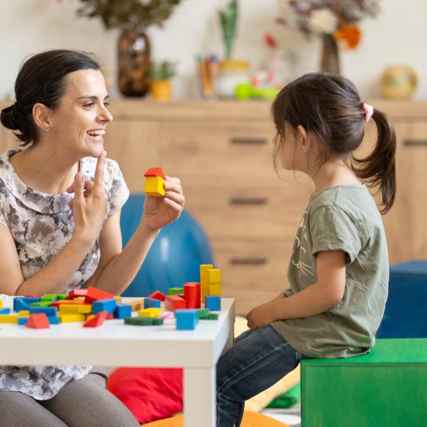Eine Frau zeigt einem Mädchen mit Bauklötzen spielerisch etwas und deutet dabei auf ihren Mund.