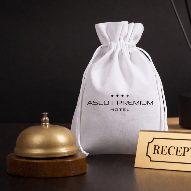 Weißer Stoffbeutel mit Hotellogo neben goldener Klingel und Rezeption-Schild auf dunklem Tisch.