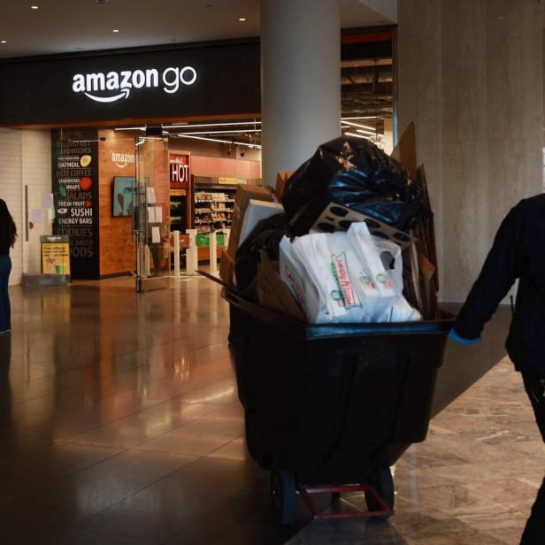 Ein Mann schiebt einen großen Müllwagen durch eine Einkaufspassage vor einem Amazon-Go-Laden.