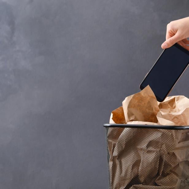Closeup view of woman throwing smartphone into trash bin on grey background, space for text. Digital detox concept