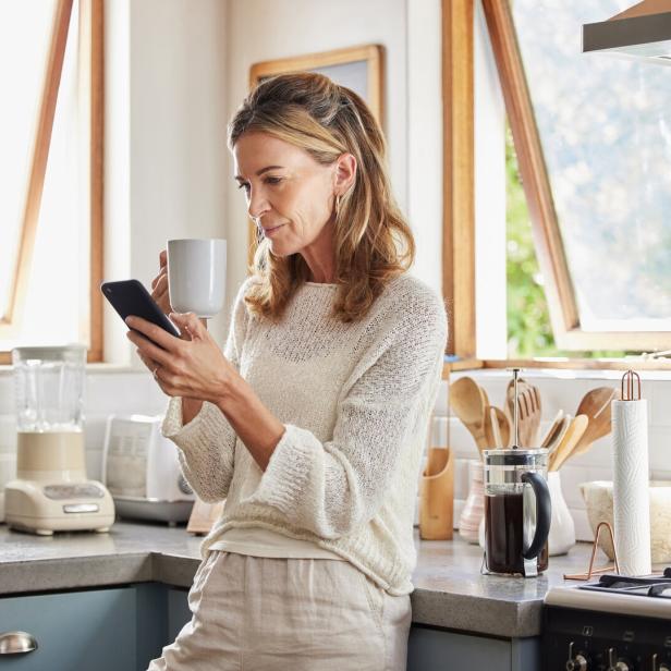 Coffee, phone and senior woman in kitchen browsing social media, text message or online app at home. Relax, tea and reading news or mobile online web surfing with female from Canada on 5g smartphone