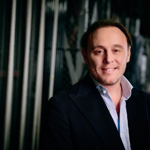 Ein lächelnder Michele Mariotti mit Sakko und hellblauem Hemd vor einem Hintergrund mit vertikalen Streben.