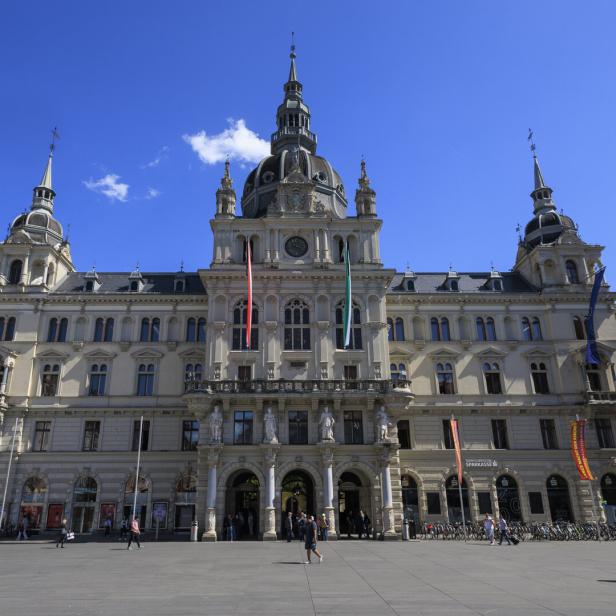 Das Grazer Rathaus präsentiert sich mit Fahnen und belebtem Vorplatz unter blauem Himmel.