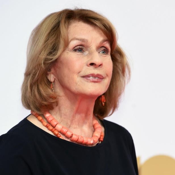 Eine Frau mit blondem Haar trägt eine korallenfarbene Halskette und Ohrringe vor einem hellen Hintergrund.