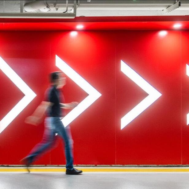 Eine Person geht vor einer roten Wand mit vier großen, leuchtenden Pfeilen nach rechts entlang.