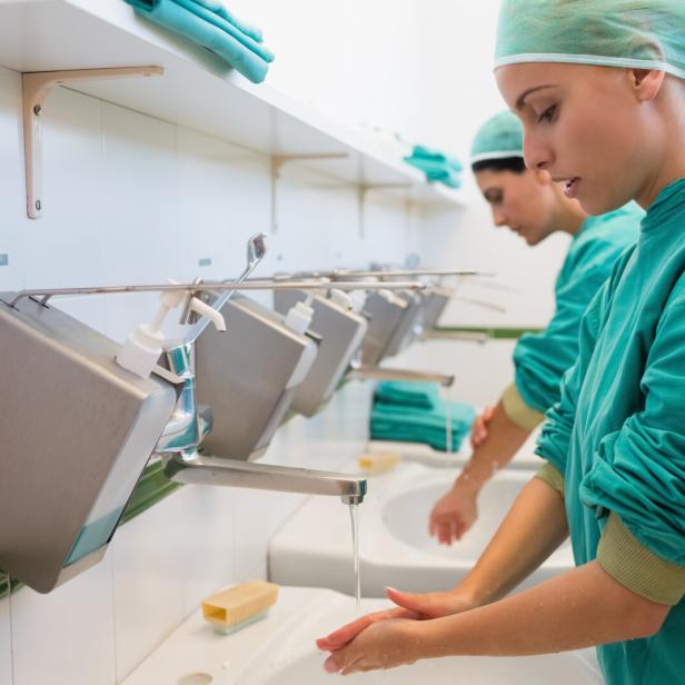 Zwei Personen in OP-Kleidung waschen sich gründlich die Hände an einem Waschbecken mit Seifenspendern.