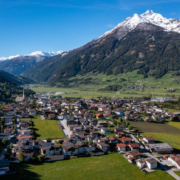 Ein Dorf liegt in einem Tal, umgeben von grünen Hängen und schneebedeckten Bergen.