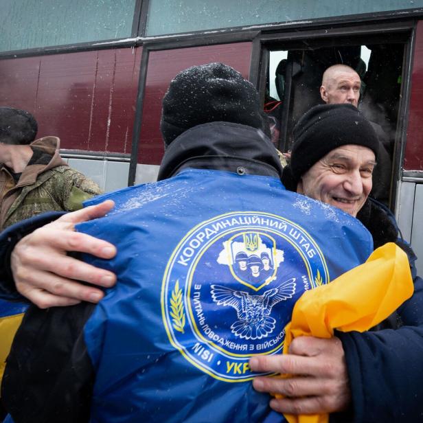 Ein Mann umarmt eine Person mit einer blauen Jacke mit ukrainischem Wappen vor einem Bus.