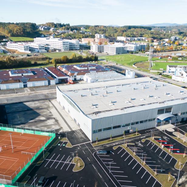 Luftaufnahme eines Gewerbegebiets mit Hallen, Tennisplätzen, Parkplätzen, Sportfeldern und Wohnhäusern im Hintergrund.