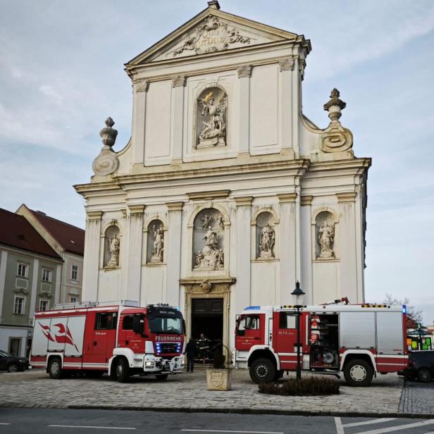 Feuerwehrfahrzeuge stehen vor der Kirche in Bruck an der Leitha, während Einsatzkräfte nach dem Brand am Eingangsbereich tätig sind.