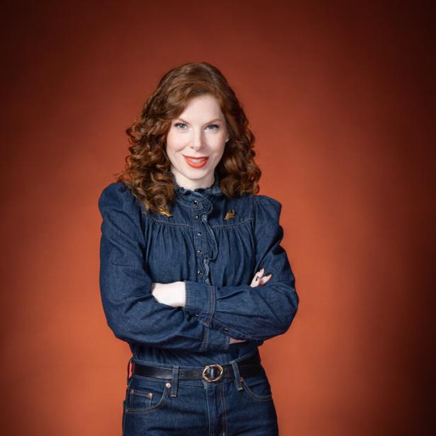 Frau mit lockigen, roten Haaren trägt eine dunkelblaue Jeansbluse und Jeanshose vor braunem Hintergrund.