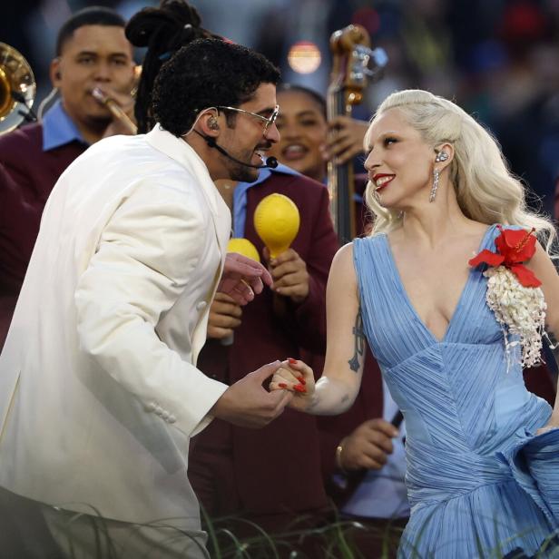 Ein Mann im weißen Anzug und eine Frau im blauen Kleid halten Händchen und lächeln sich auf einer Bühne mit Musikern an.