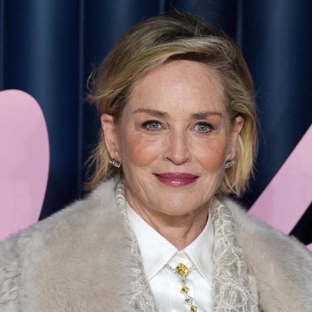 Sharon Stone mit kurzem blonden Haar, Ohrringen und einer Brosche lächelt.