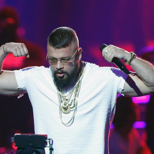 Ein Mann mit Bart und Goldketten zeigt seine Muskeln auf einer Bühne mit buntem Licht.