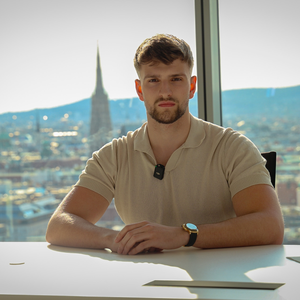 Kilian Kropiunik sitzt an einem Schreibtisch vor der Wiener Skyline und blickt ernst in die Kamera.