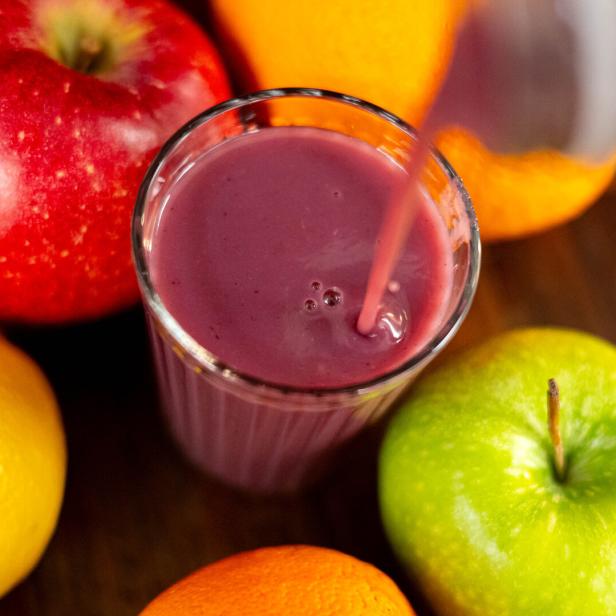 Ein Glas lila Smoothie wird umgeben von Apfel, Orange und Zitrone eingeschenkt.