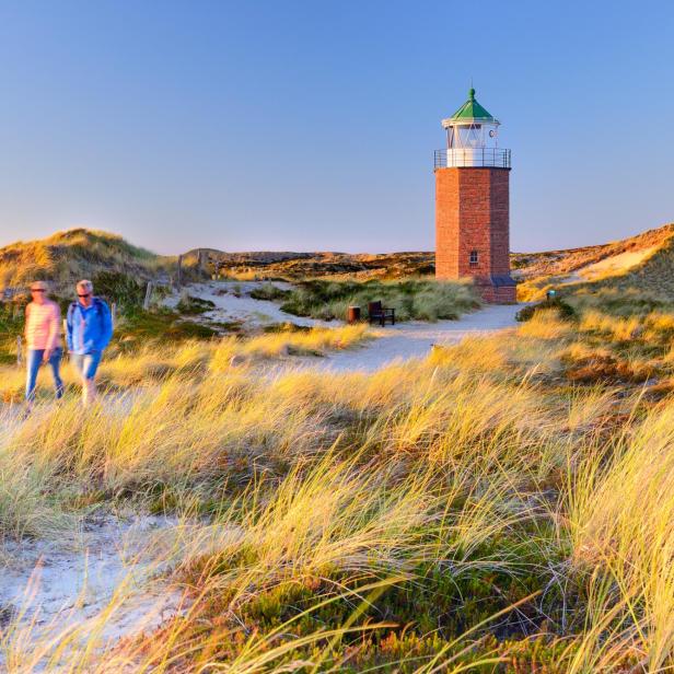 Zwei Personen gehen auf einem sandigen Weg durch Dünen, im Hintergrund steht ein roter Leuchtturm mit grünem Dach.