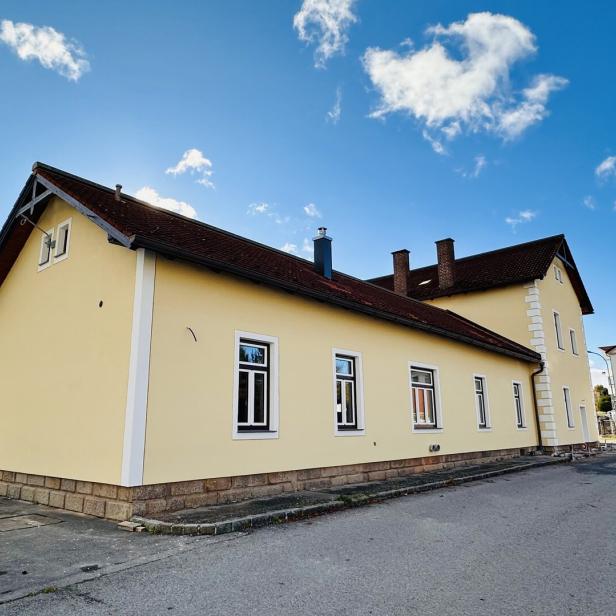 Eine Außenaufnahme vom beigen Bahnhofsgebäude vor einem blauen, fast wolkenlosen Himmel.