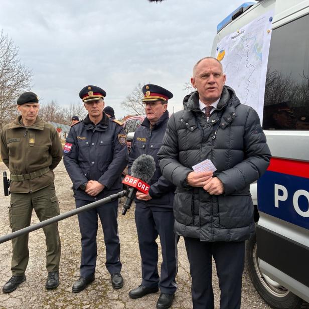 Ein Mann in Zivilkleidung spricht vor Mikrofonen, flankiert von Polizisten vor einem Einsatzfahrzeug mit Karten.