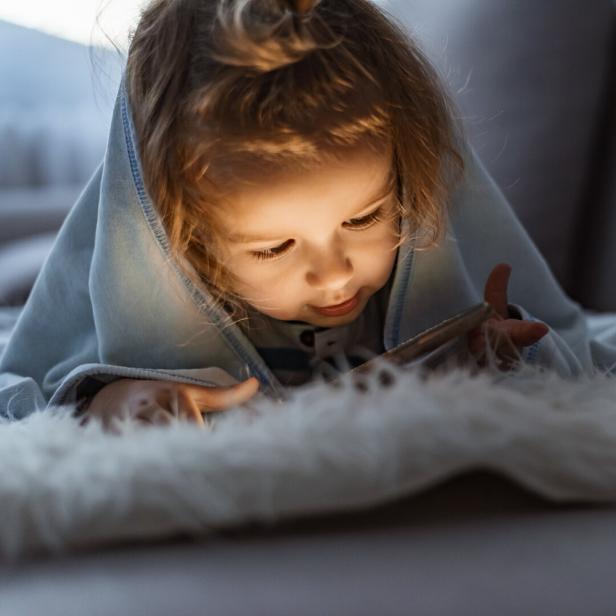 Ein kleines Mädchen liegt in eine Decke gehüllt und schaut auf ein leuchtendes Handy.