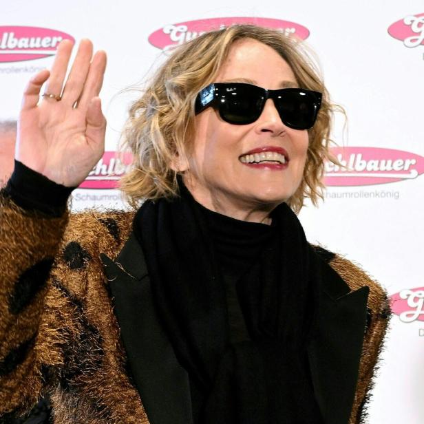 Eine Frau mit Sonnenbrille und lockigem Haar lächelt und winkt, im Hintergrund sind Werbeschilder zu sehen.