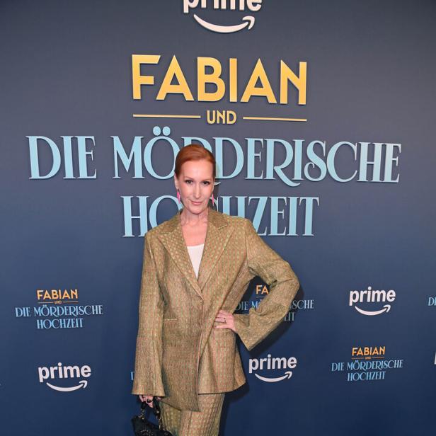 Eine Frau mit rotem Haar (Nicole Beutler) trägt einen gemusterten Hosenanzug und pinke Schuhe, steht vor einem blauen Hintergrund mit Schriftzug.